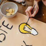 A close-up of a person painting a light bulb design on a poster with a paintbrush.